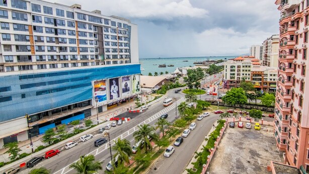 A multi-story hotel with a large billboard, a busy street with cars, and a view of the sea. - Marina Travellers Suite Marina Court (Kota Kinabalu)