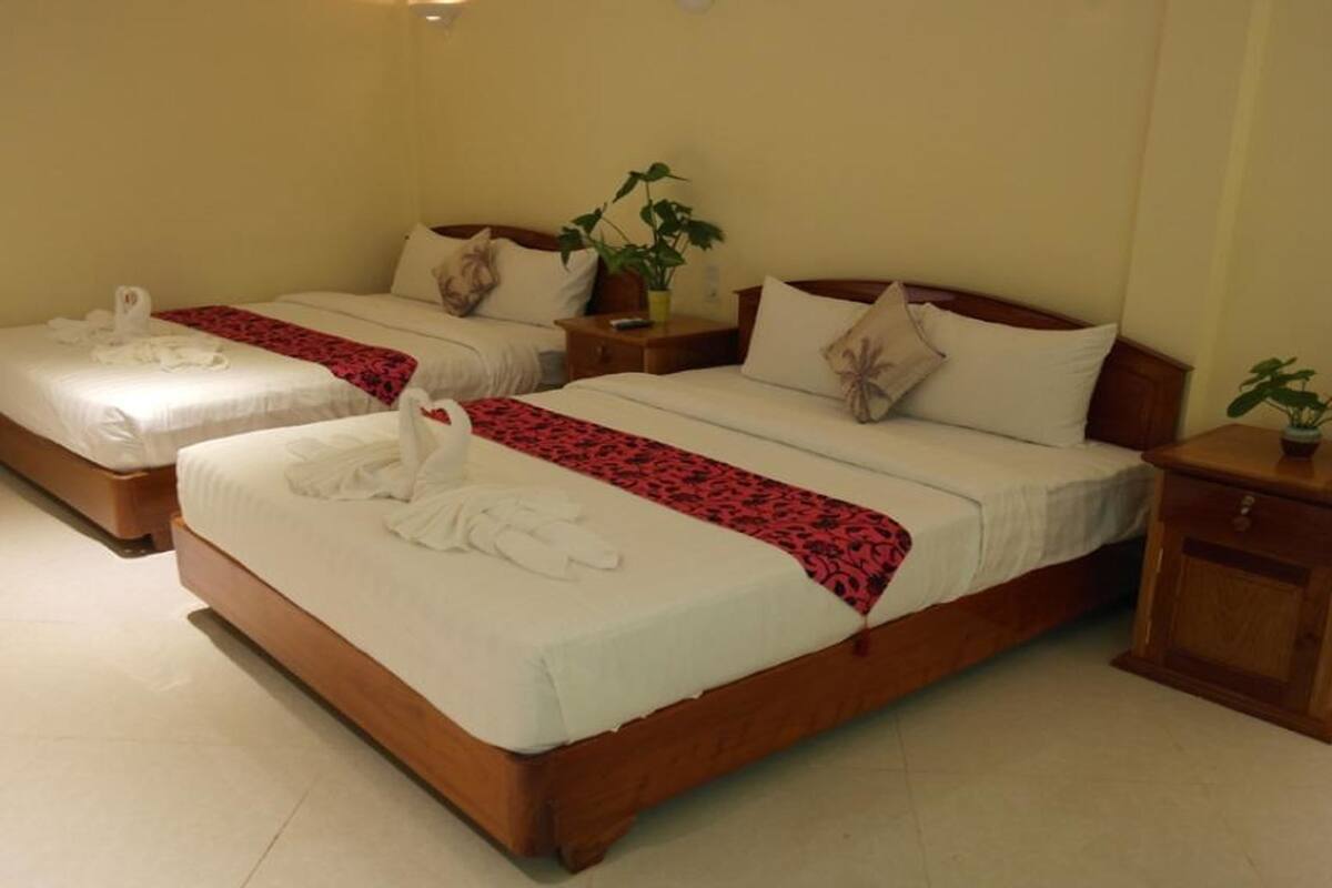Habitación de hotel con dos camas individuales, cada una con ropa blanca, un corredor estampado rojo y un par de toallas blancas. Incluye cabeceros y mesitas de noche de madera con plantas en macetas.