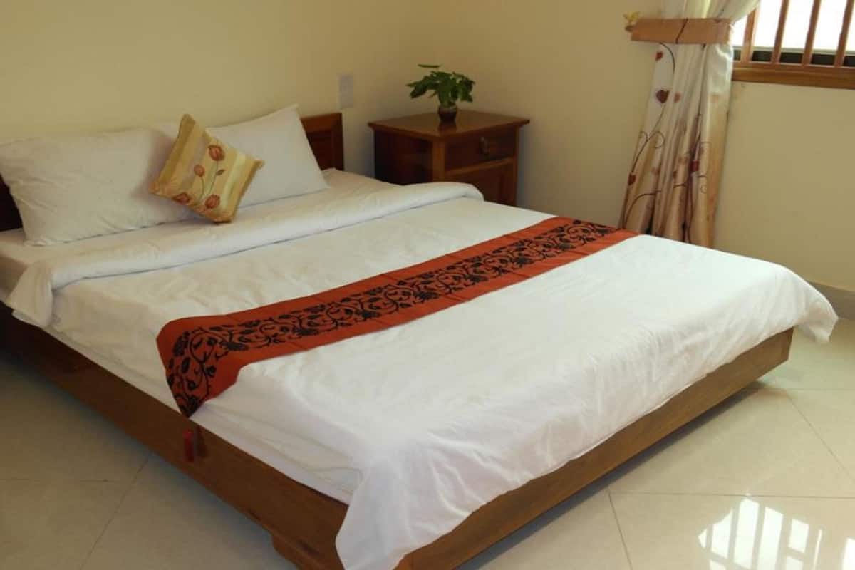 Una cama bien hecha con sábanas blancas y una manta roja estampada, una mesita de noche con una planta y una ventana con cortinas.