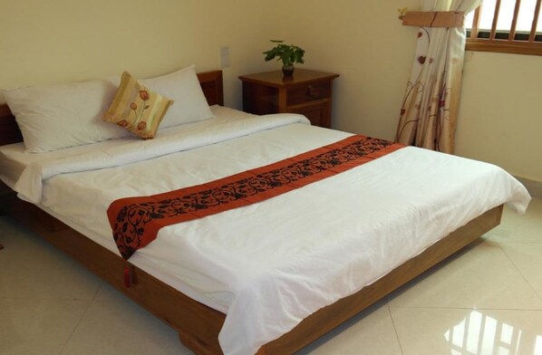 Una cama bien hecha con sábanas blancas y una manta roja estampada, una mesita de noche con una planta y una ventana con cortinas.