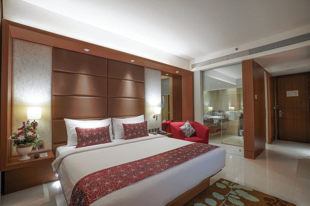 A hotel room with a large bed, a red armchair, a wooden headboard, and a glass shower area.