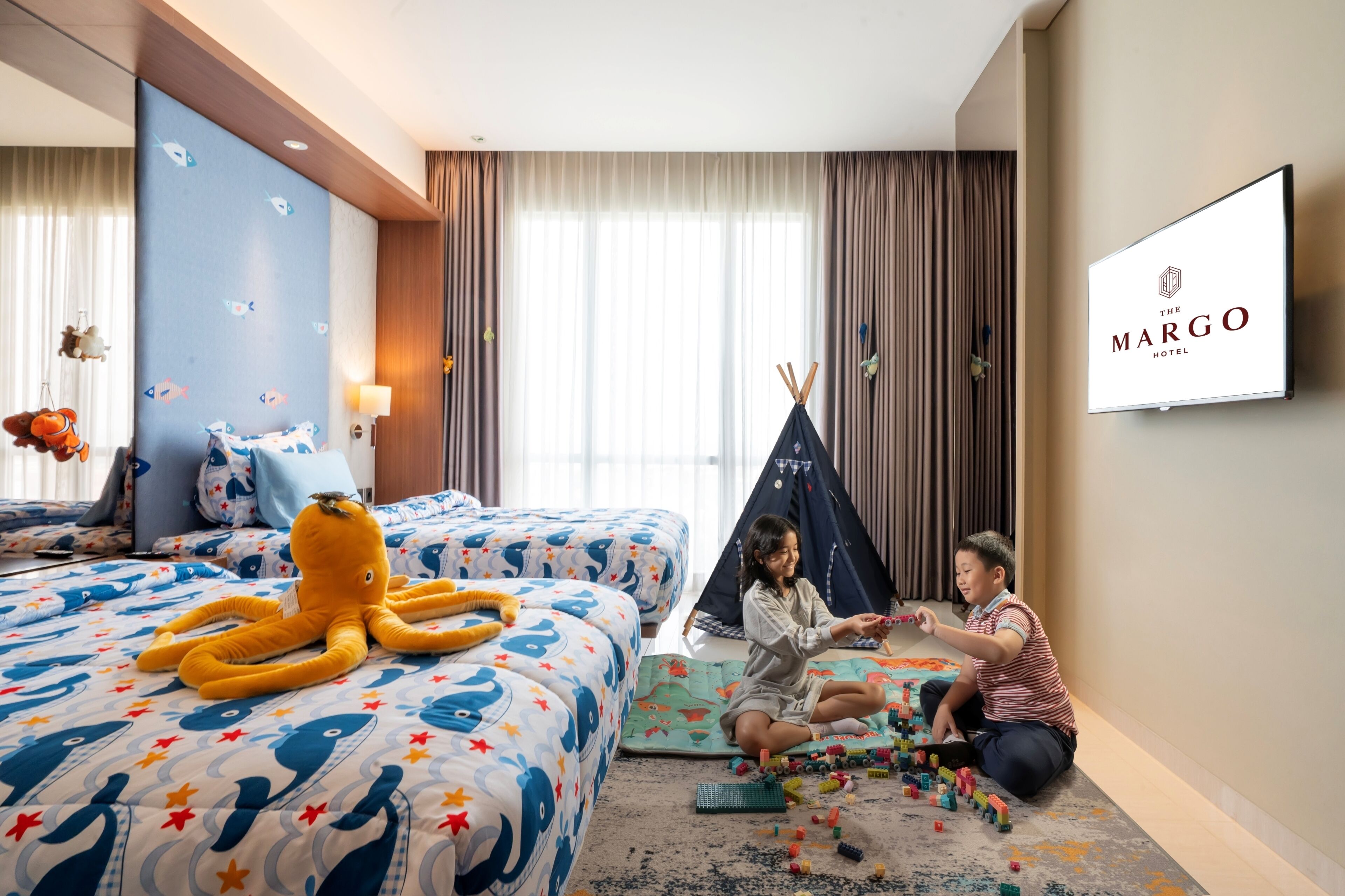 A hotel room with a bed, a child playing with toys, a tent, and a television displaying the hotel name.