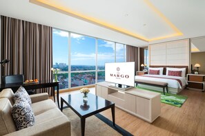 A hotel room with a bed, a TV, a sofa, a desk, and a large window with a city view. - The Margo Hotel (Depok)