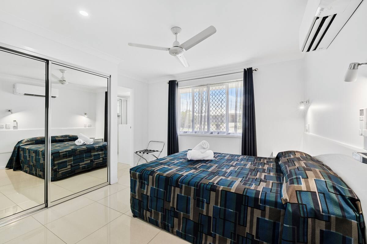 A modern hotel room with a large bed, a ceiling fan, a window with curtains, and a bathroom with a mirror and towel rack.