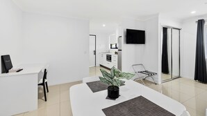 A modern, well-lit room with a dining table, chairs, a small kitchen area, a TV mounted on the wall, and a sliding glass door leading to a balcony. - Casa Nostra Motel (Parkhurst)