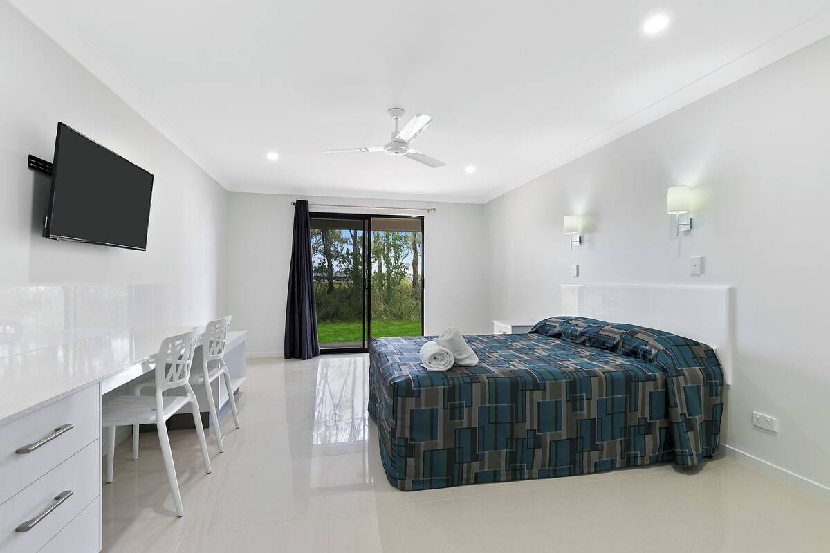 A modern hotel room with a flat-screen TV, a bed with a patterned bedspread, a desk with chairs, and a large sliding glass door leading to a balcony.