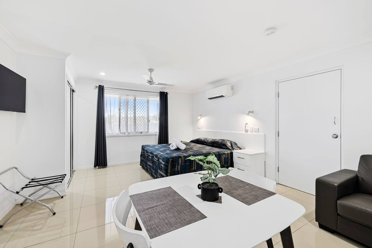 A modern hotel room with a bed, a dining table with placemats, a plant, a ceiling fan, and a TV mounted on the wall.