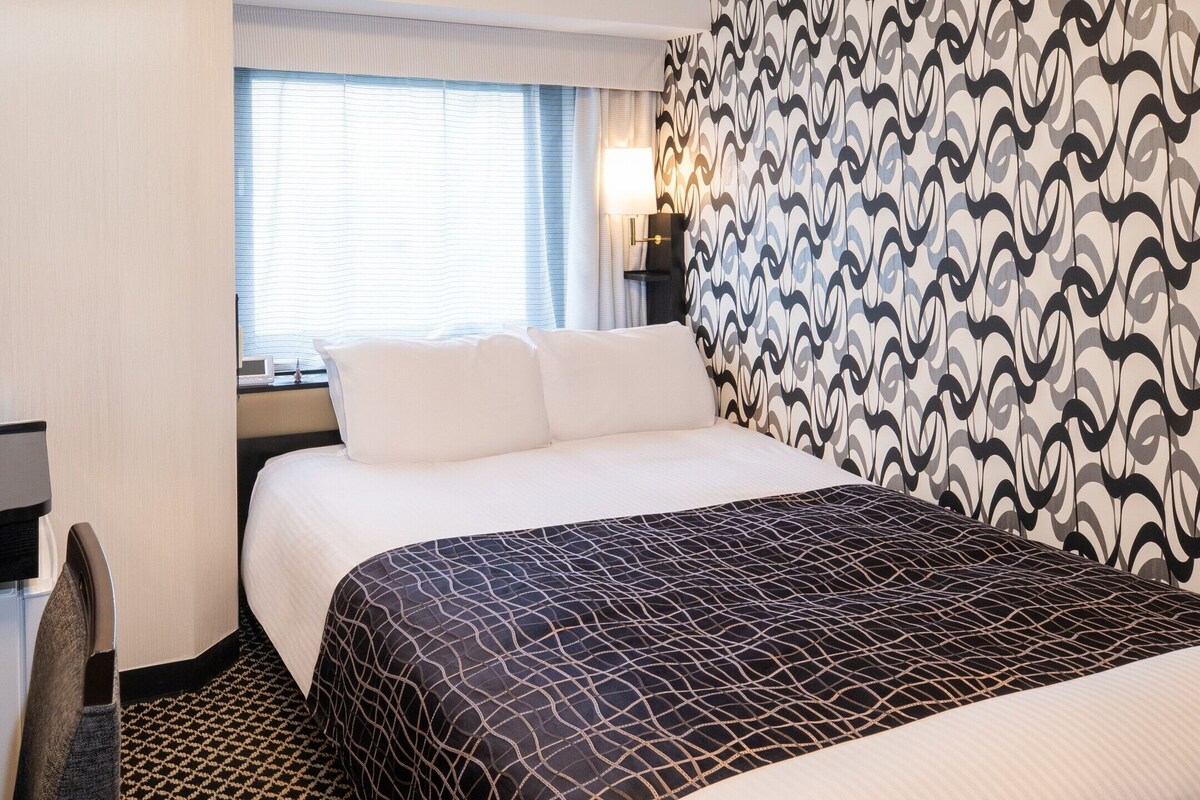 A hotel room with a bed, a desk, a telephone, a window with blinds, and patterned curtains.