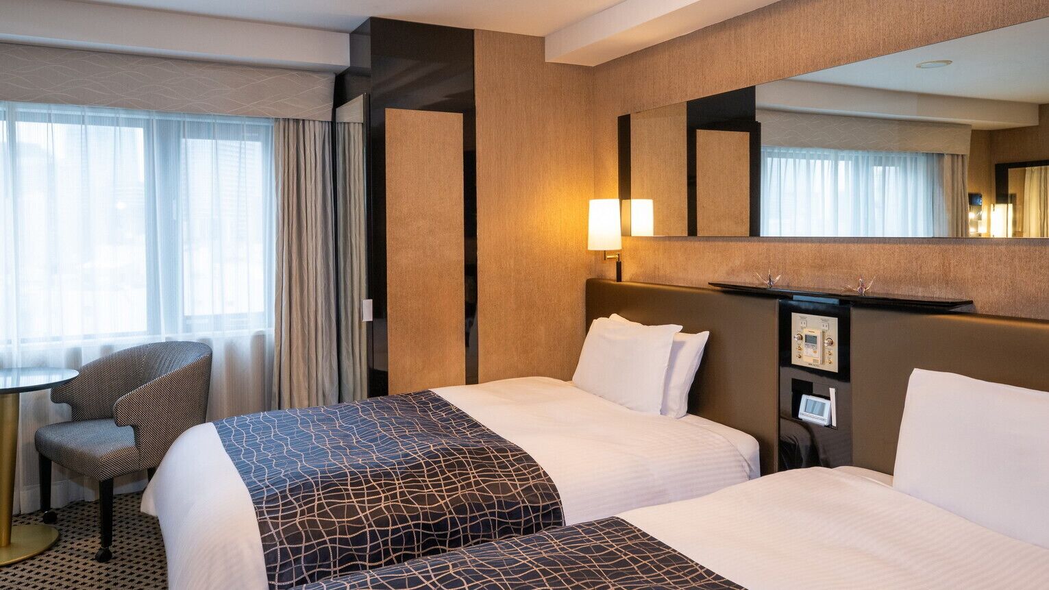 A hotel room with two beds, a desk, a chair, a large mirror, and a wall-mounted control panel.