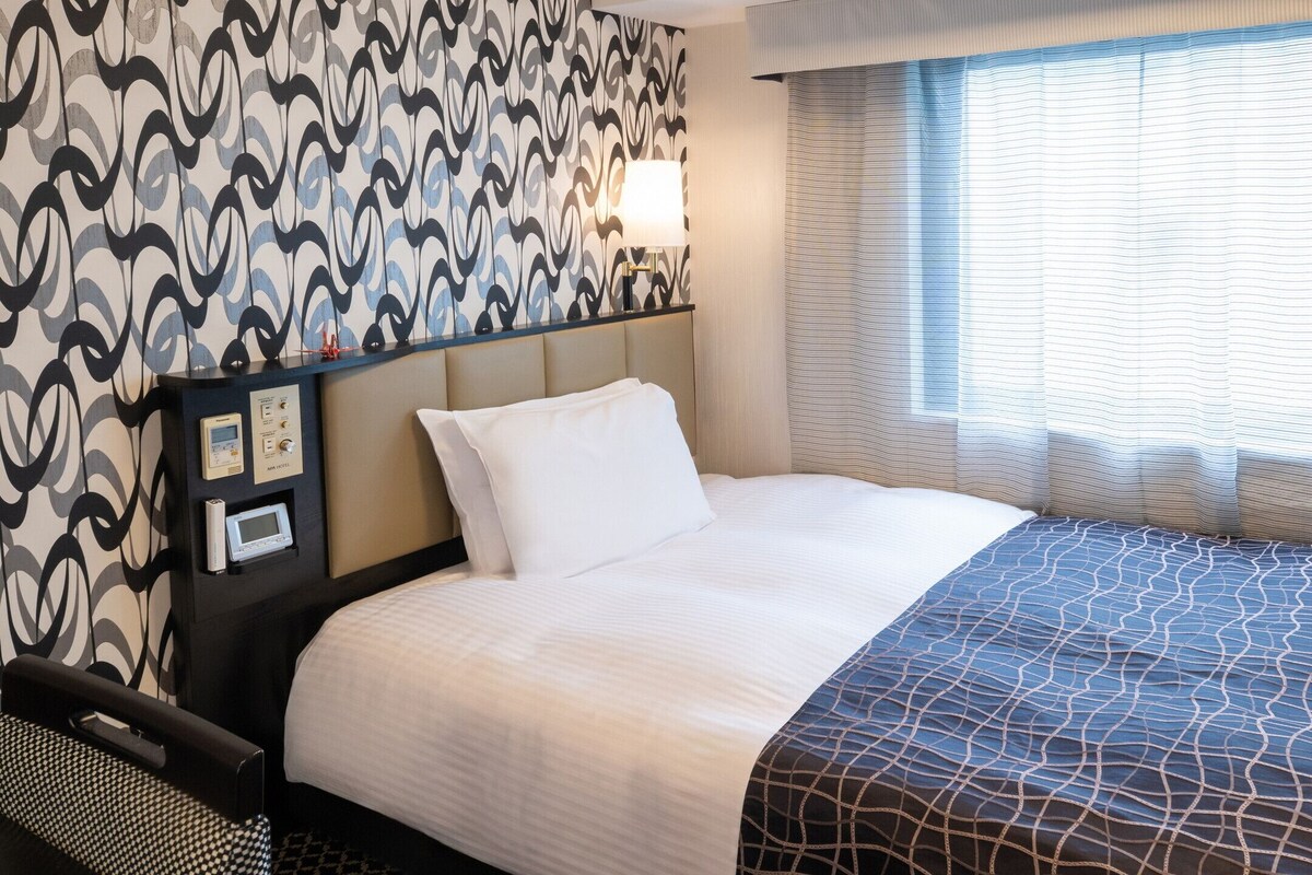 A hotel room with a bed, a control panel, a window with curtains, and a patterned wall.