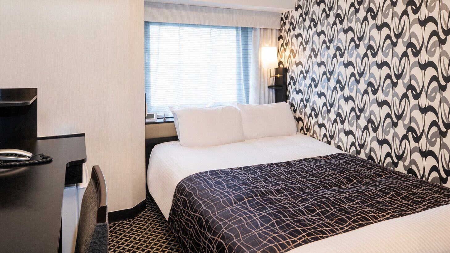 A hotel room with a bed, a desk, and a window with blinds.