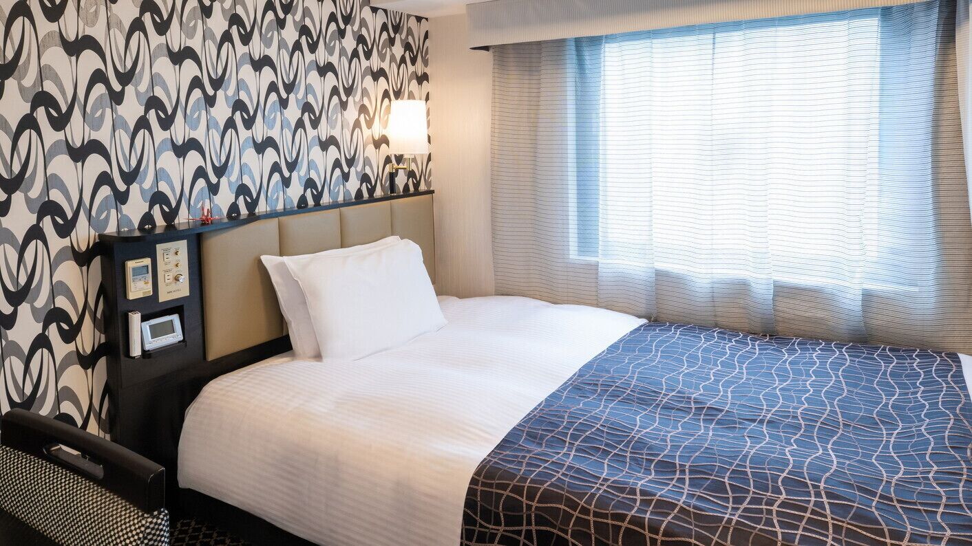A hotel room with a bed, a headboard, a bedside table with a lamp, a window with curtains, and a wall with a patterned wallpaper.