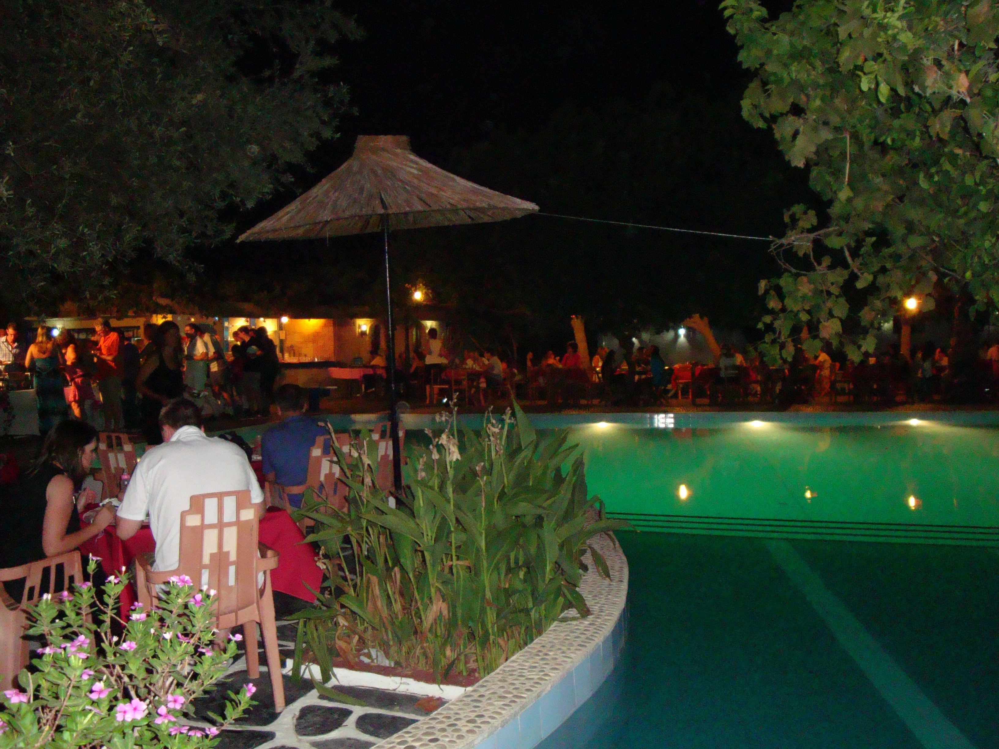 An outdoor pool area with a thatched umbrella, seating, and a dining area with people.