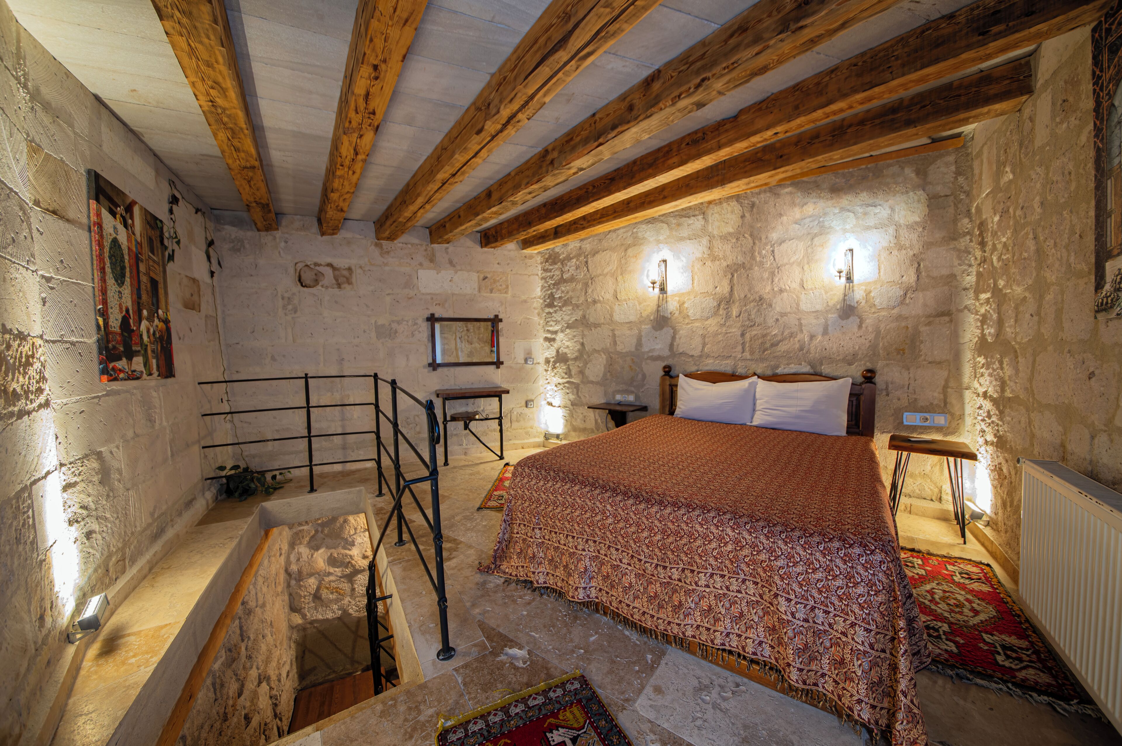 A rustic bedroom with exposed wooden beams, a stone wall, a bed with a patterned blanket, a small table, and a wall-mounted light fixture.