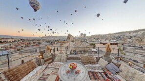 A rooftop terrace with a view of a cityscape, a table with a fruit bowl and glasses, and a variety of cushions and rugs. - Chelebi Cave House (Nevsehir)