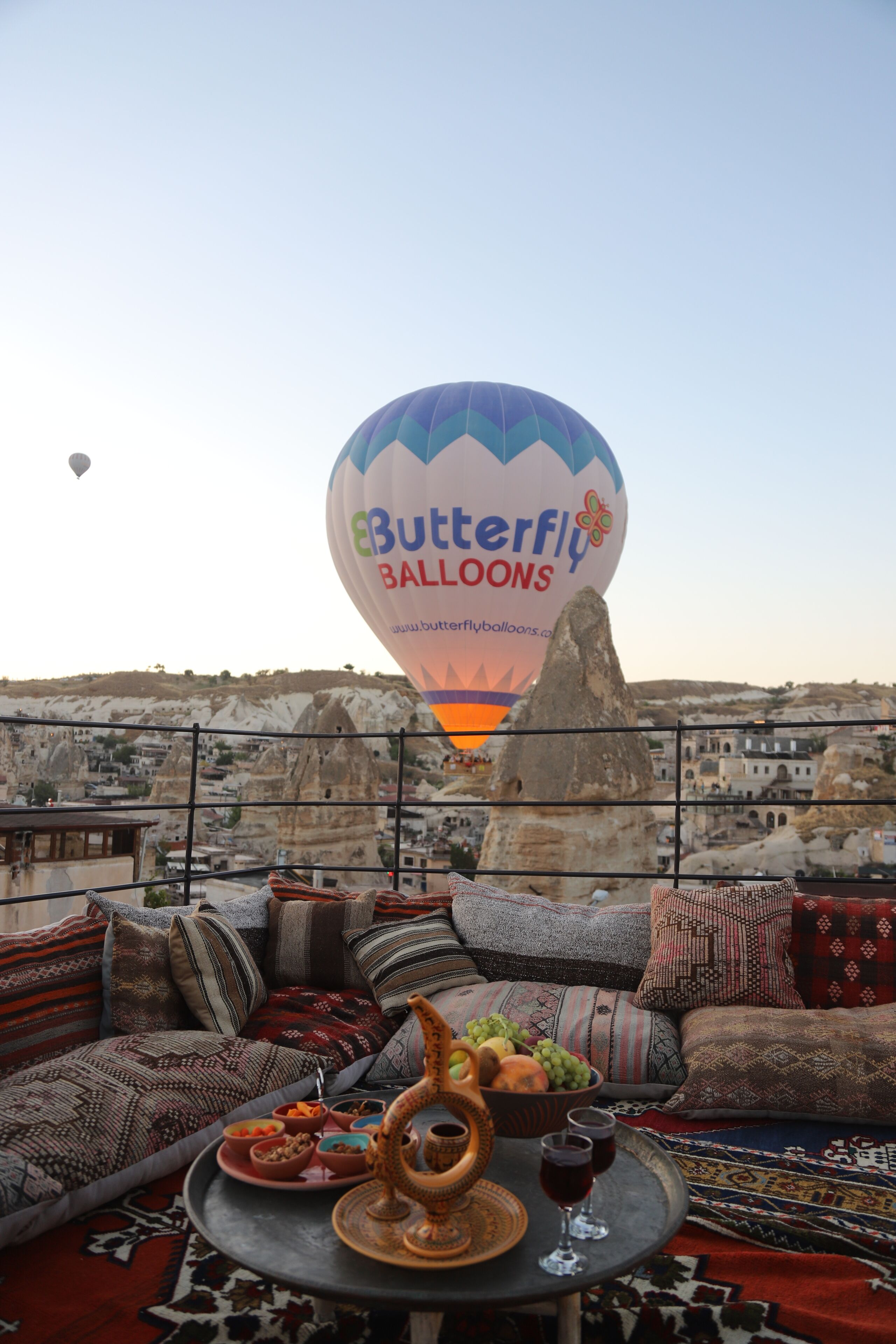 A rooftop terrace with a view of a cityscape, a hot air balloon, and a table with a traditional drink set.