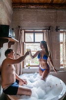 Un hombre y una mujer en una bañera llena de burbujas, con una ventana y una botella en el alféizar.