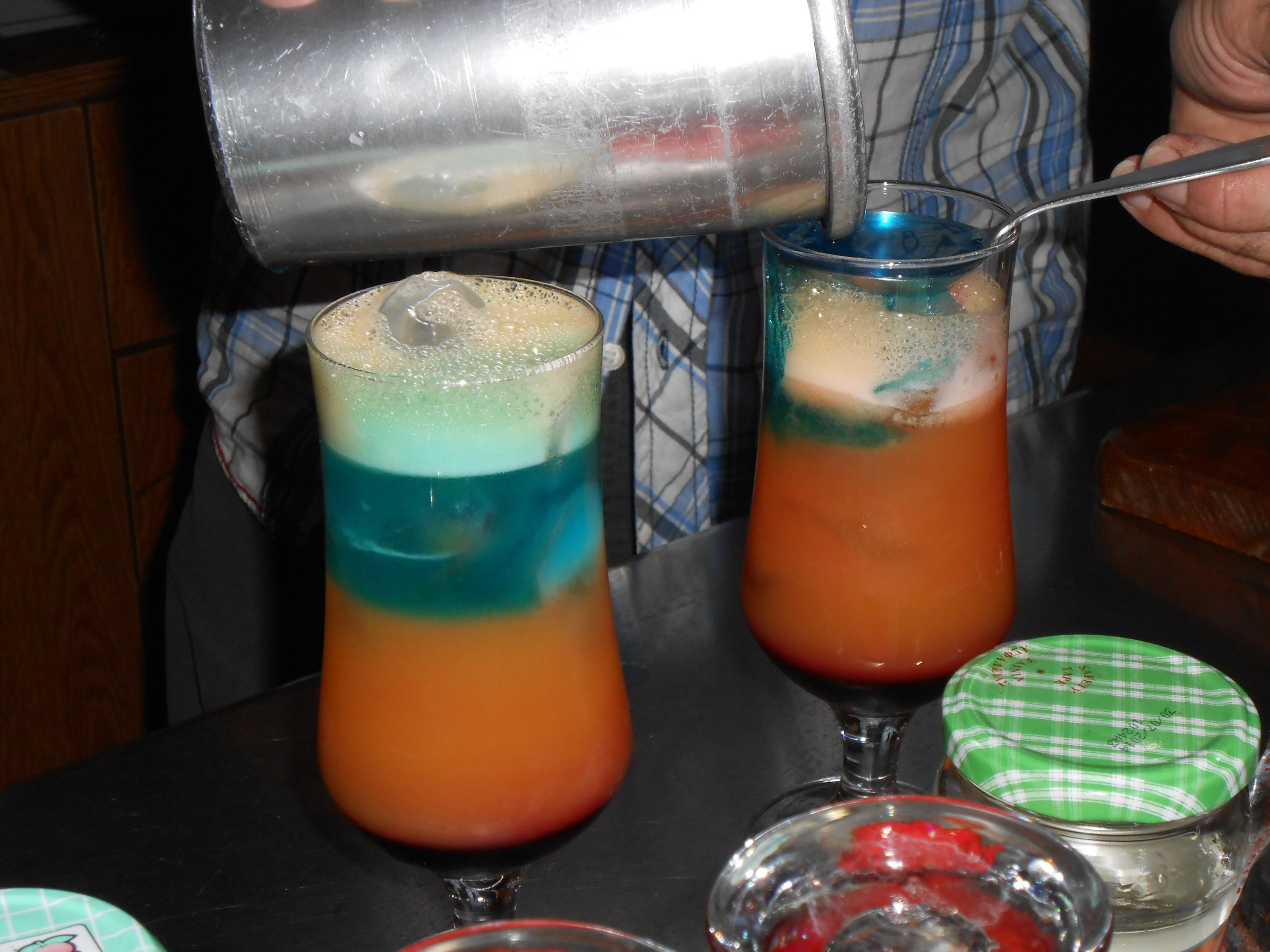 Two cocktails in tall glasses with layered colors, a metal shaker, and a spoon.