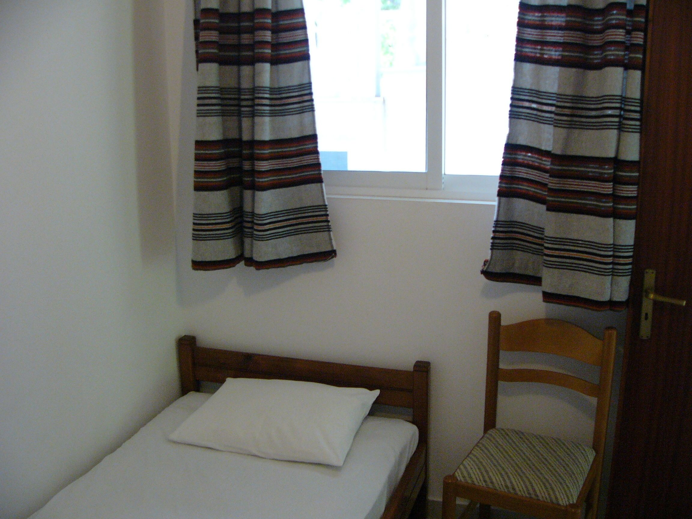 Ein kleines, schlichtes Zimmer mit einem Bett, einem Holzstuhl und zwei Vorhängen.