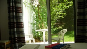 Una habitación con una cama, una mesita y una silla. Se ve vegetación por la ventana.