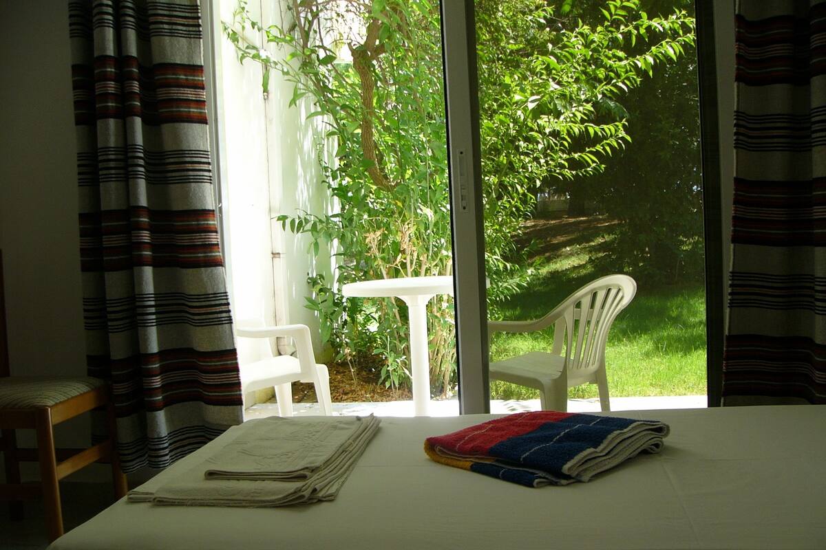 A room with a bed, a small table, and a chair. There is a window with a view of greenery outside.