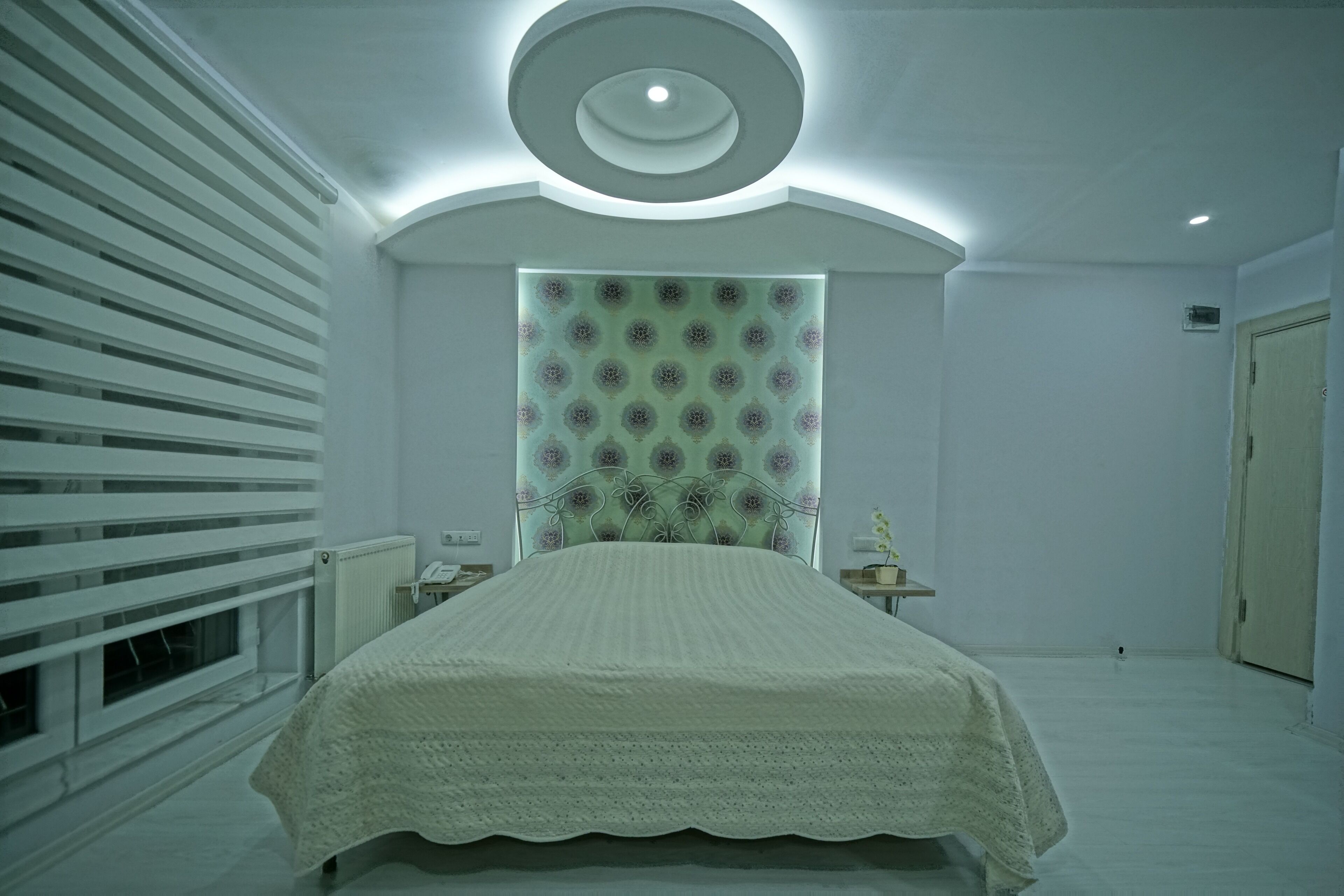 A bedroom with a bed, a radiator, a window with blinds, a wall-mounted light fixture, and a door.