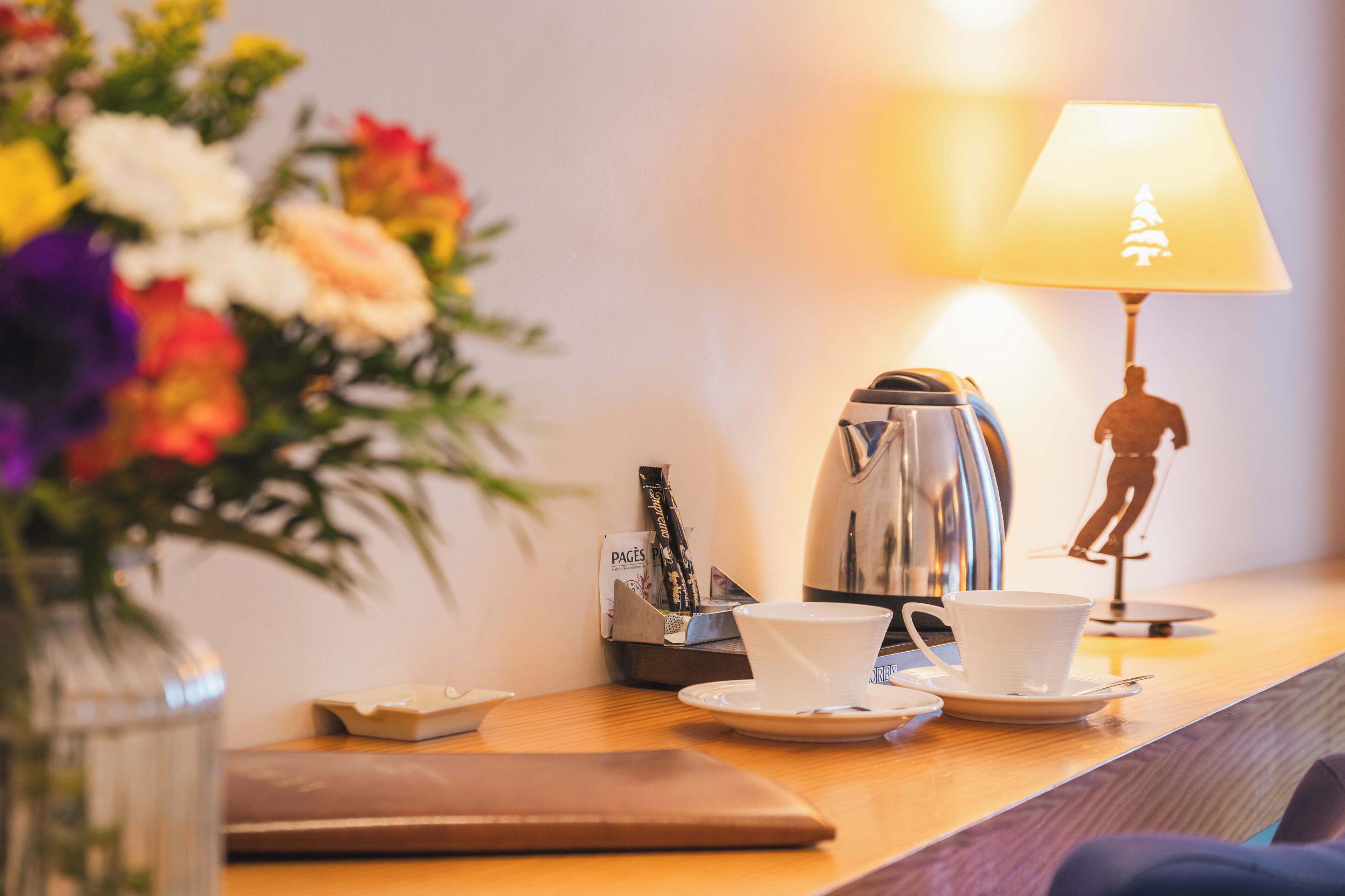 Une table en bois, avec une machine à café, une tasse, une soucoupe, une lampe décorée d’un motif de sapin de Noël et un vase rempli de fleurs.