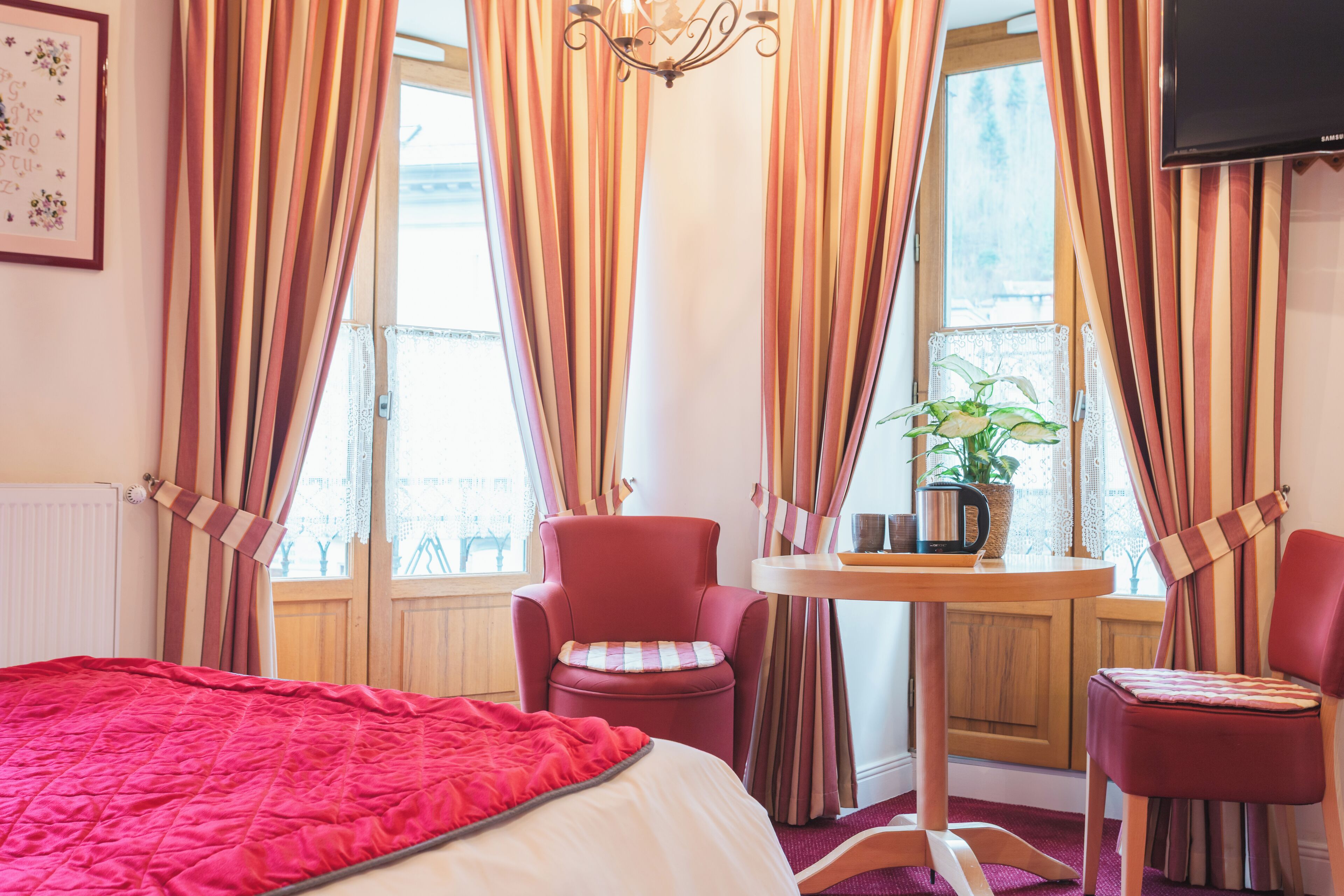Ett hotellrum med en säng, två stolar, ett litet bord med en växt och gardiner.