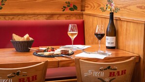 Interior de madera con un banco tapizado rojo, una mesa con una botella de vino, copas, pan y una cesta de pan.