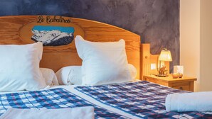 Una habitación de hotel con cabecera de madera, edredón acolchado, almohadas blancas, mesita de noche con lámpara y un cuadro en la pared que representa montañas.
