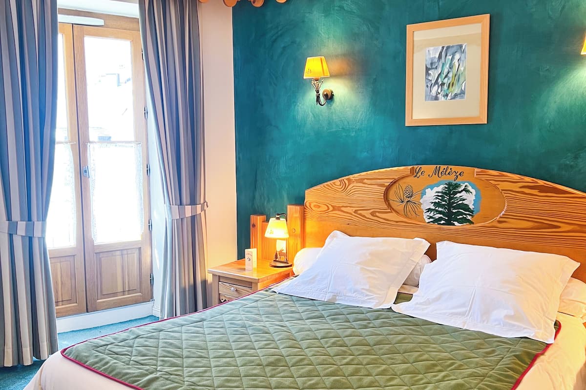 Une chambre d’hôtel avec un grand lit, une tête de lit en bois, une table de chevet avec une lampe, un tableau encadré au mur et une vue sur l’extérieur par une fenêtre avec des rideaux.