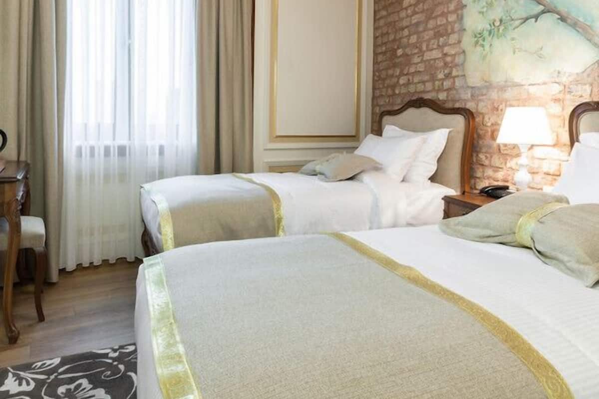 Habitación de hotel con dos camas, un ventanal con cortinas, una pared de ladrillo y un cuadro de un árbol.
