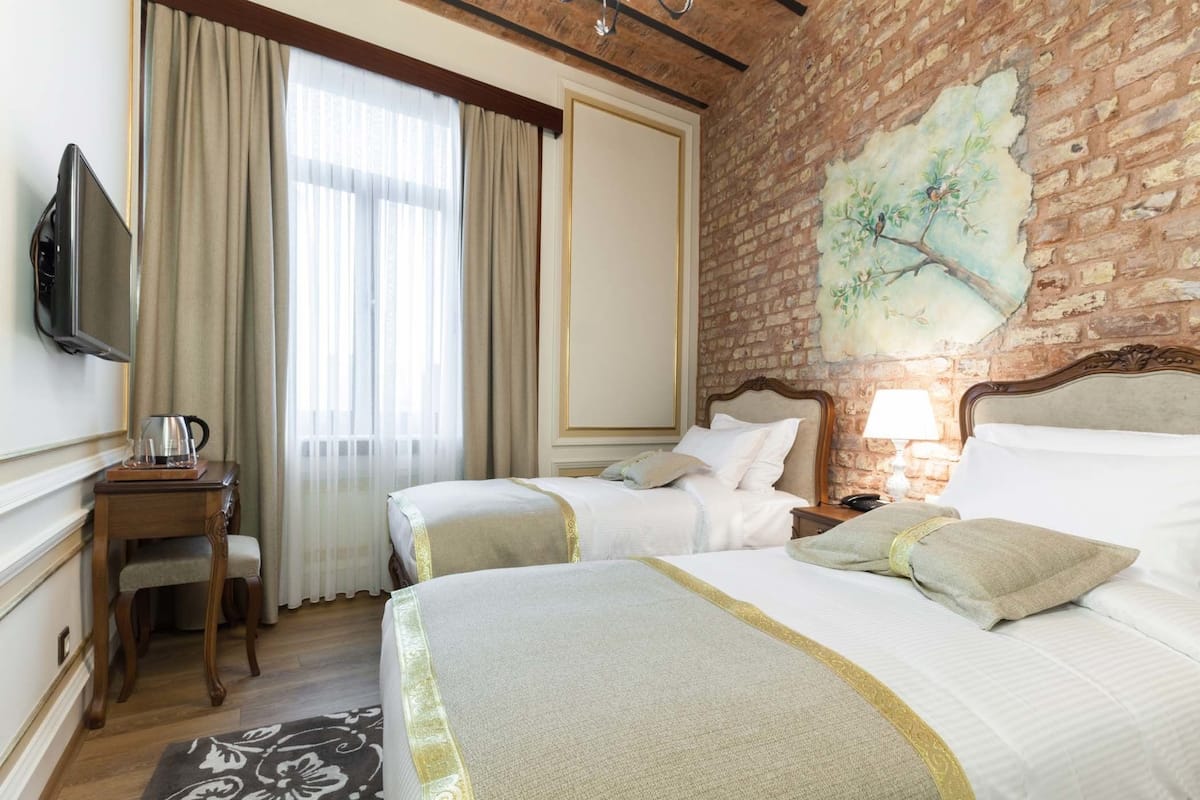 Habitación de hotel con dos camas, un escritorio con lámpara, televisor y pared de ladrillo con mural.