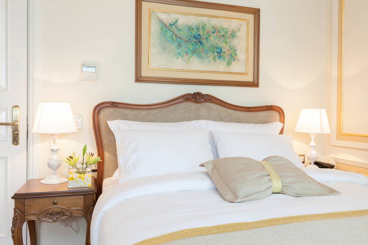 Una cama bien hecha con sábanas blancas y un cojín decorativo, flanqueada por dos mesitas de noche con lámparas y un florero.