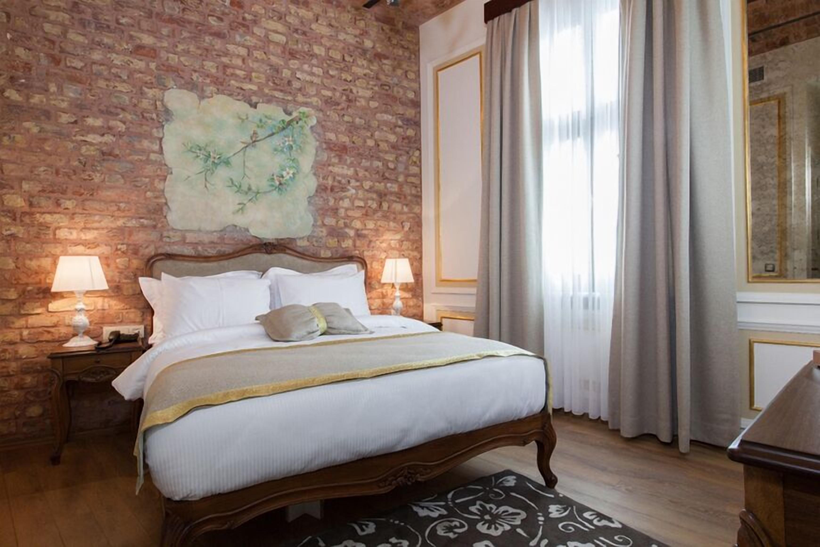 Un dormitorio con pared de ladrillo, una cama con ropa de cama blanca, dos lámparas de noche y un ventanal con cortinas.
