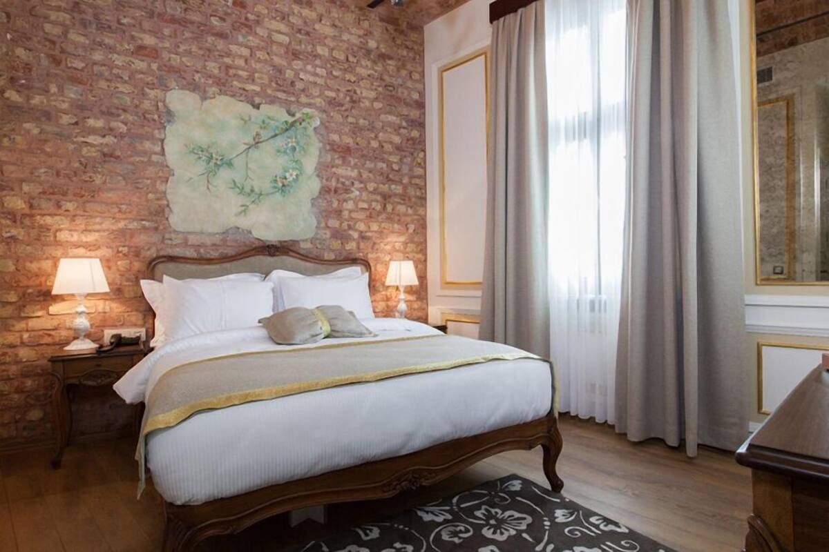 A bedroom with a brick wall, a bed with white bedding, two bedside lamps, and a large window with curtains.