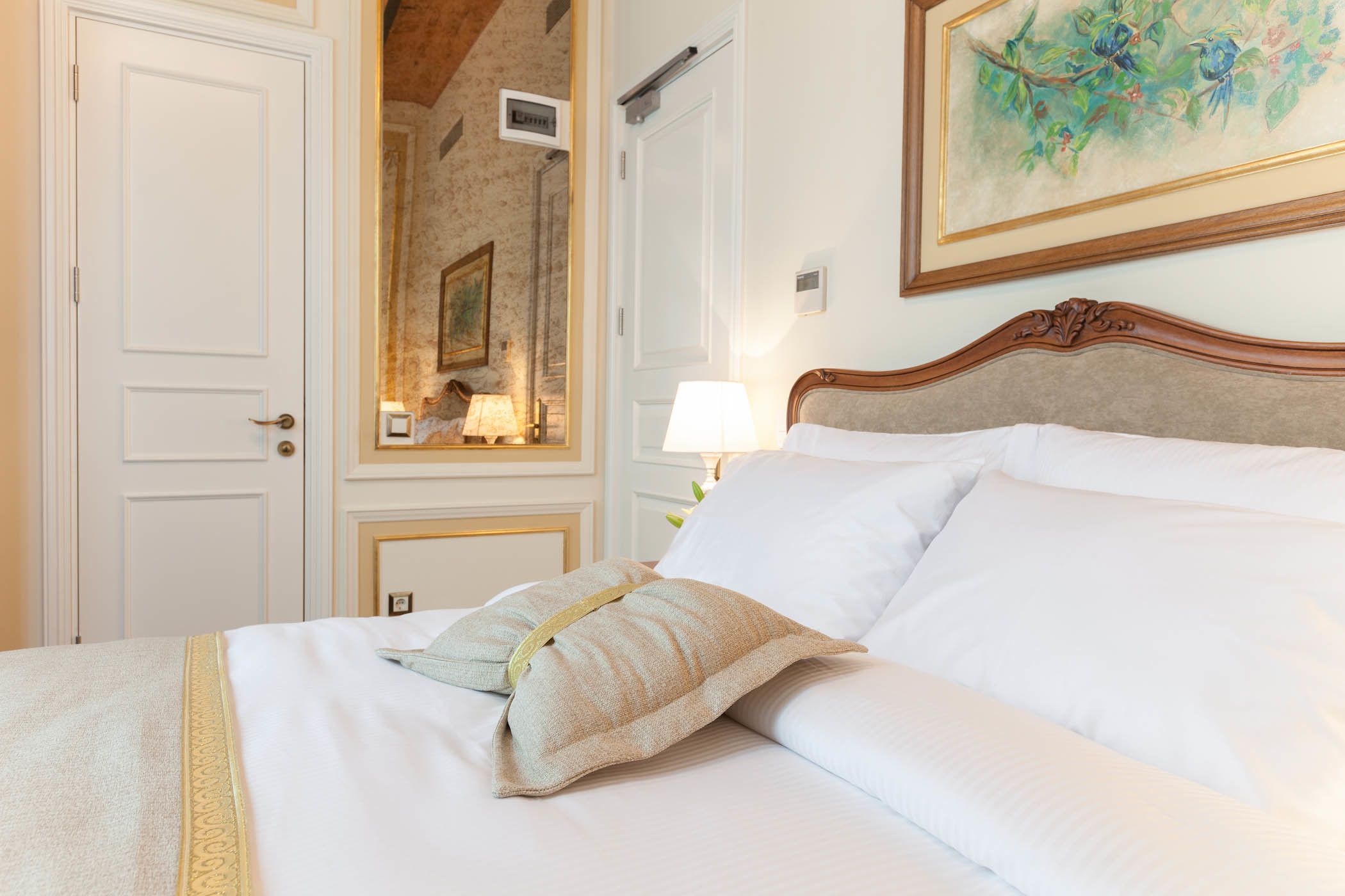 A neatly made bed with white linens and two pillows, a wooden headboard, a nightstand with a lamp, and a framed painting on the wall.