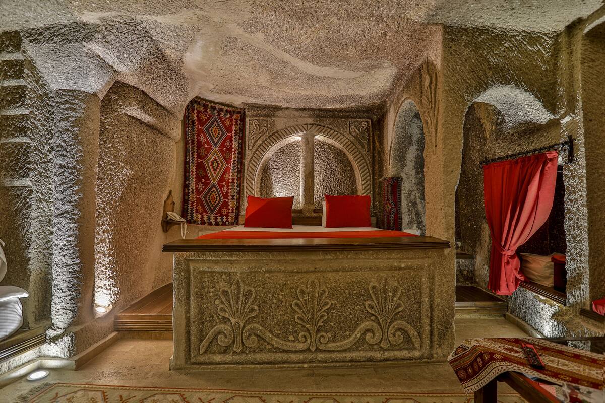 Quarto com paredes de pedra, uma cama, uma cortina vermelha e um tapete estampado.