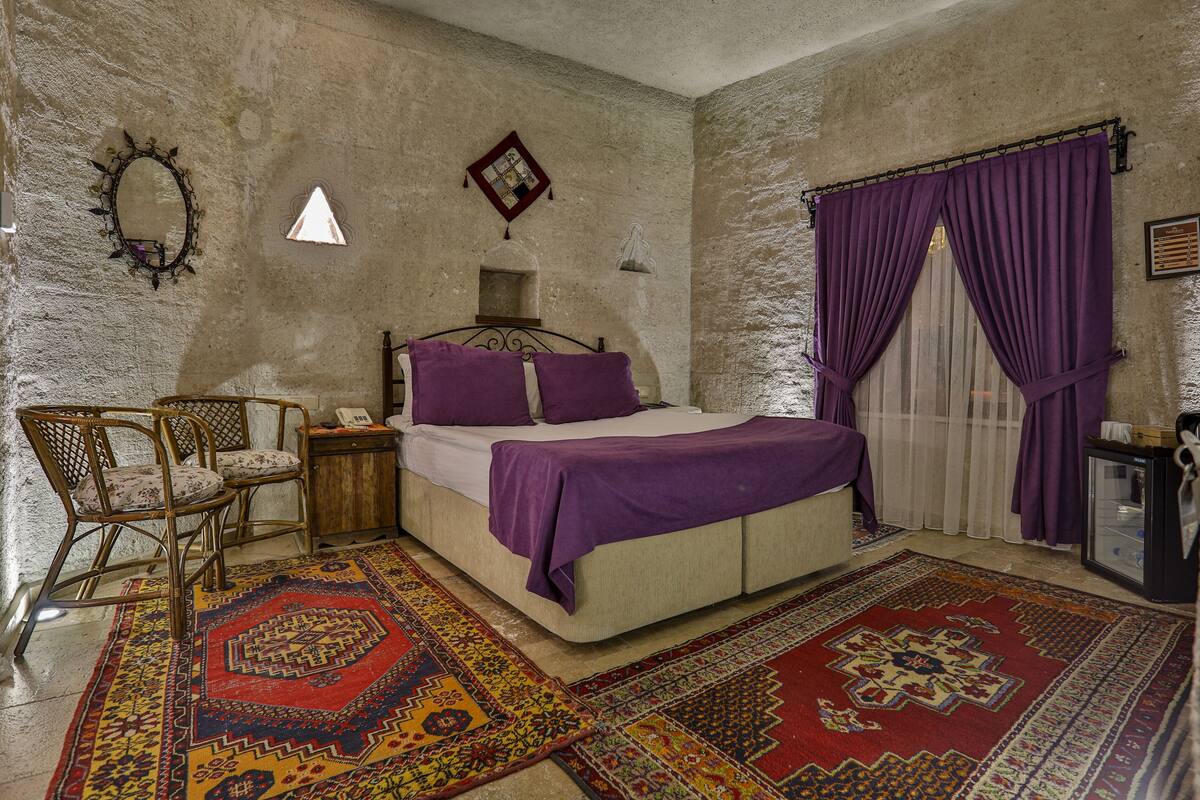 Quarto com cama, cadeira, mesa de cabeceira, espelho e janela com cortinas roxas.