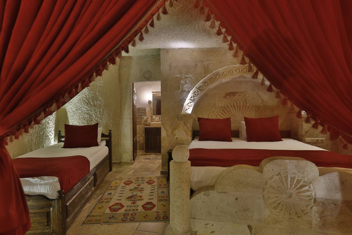 Quarto com duas camas, cada uma com roupa de cama vermelha, um tapete estampado e uma parede de pedra com detalhes entalhados.