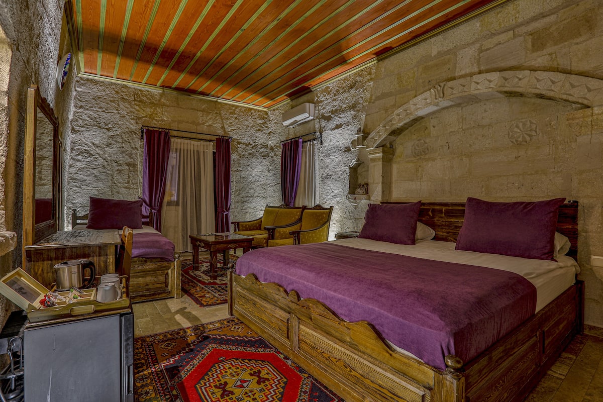 Quarto tradicional com parede de pedra, cama de madeira e pequena escrivaninha com espelho.