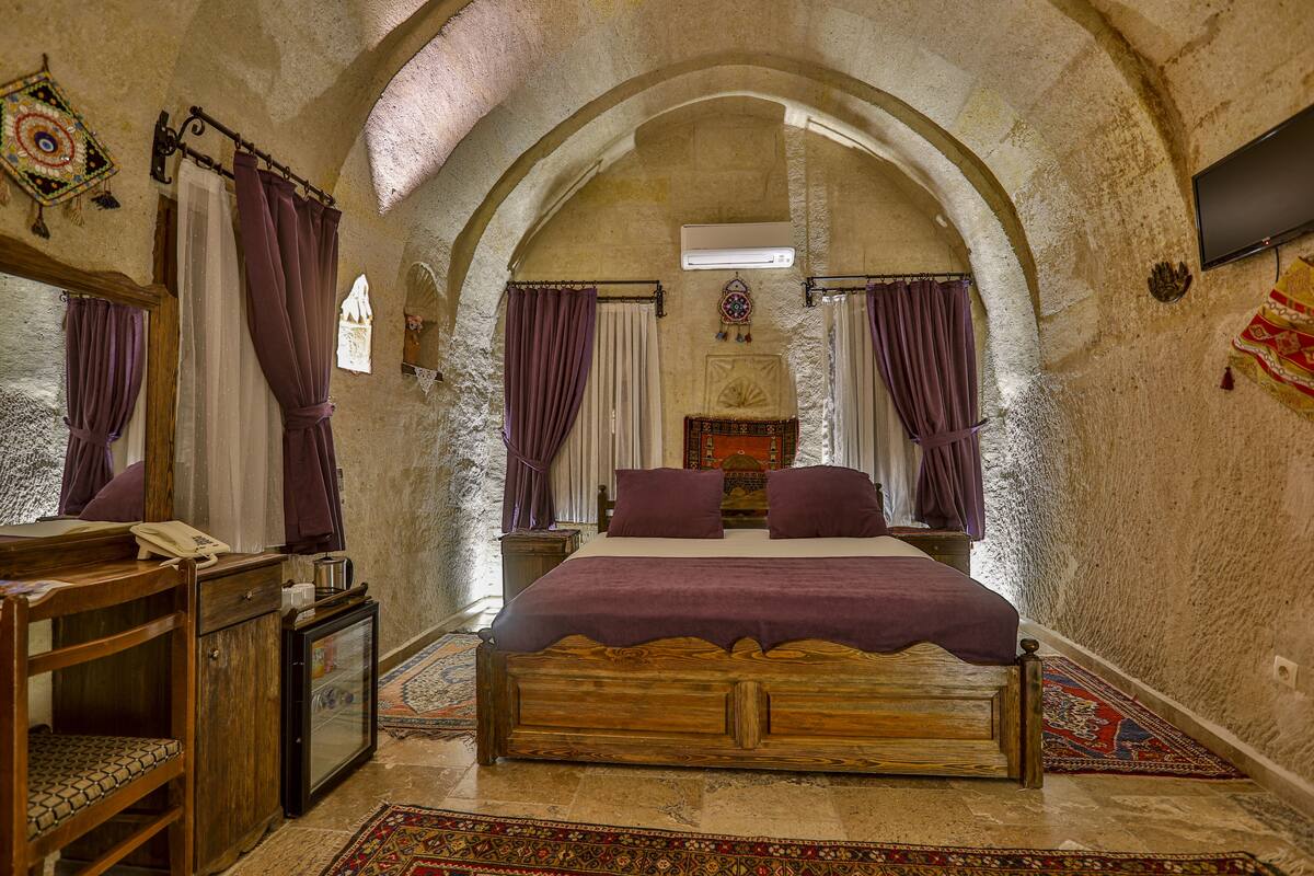 Quarto com cama de madeira, cortinas roxas, uma mesinha de cabeceira e televisão na parede.