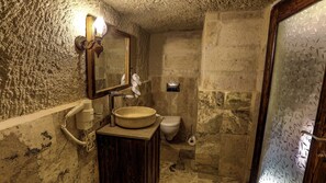 A bathroom with a stone wall, a wooden vanity, a round sink, a toilet, and a shower area. - Hidden Cave (Nevsehir)