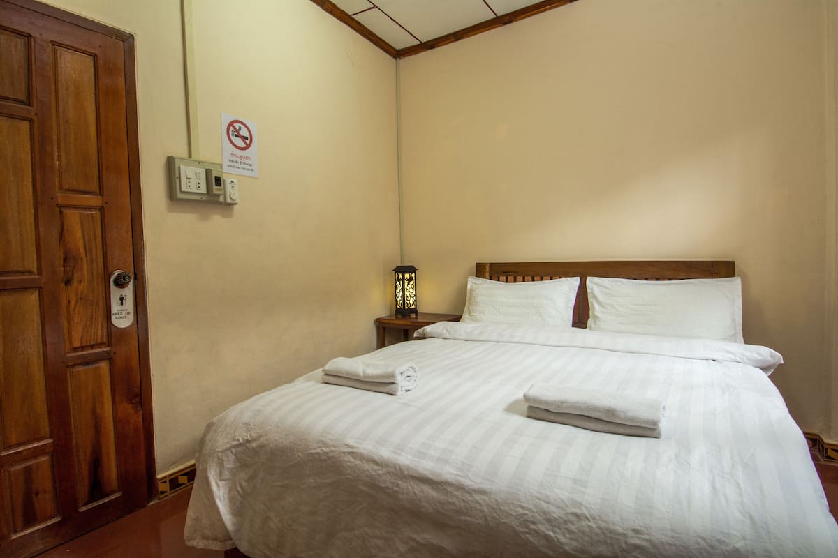Une chambre d’hôtel avec une porte en bois, un lit avec du linge de maison blanc, une table de chevet avec une lampe et un panneau indiquant qu’il est interdit de fumer.