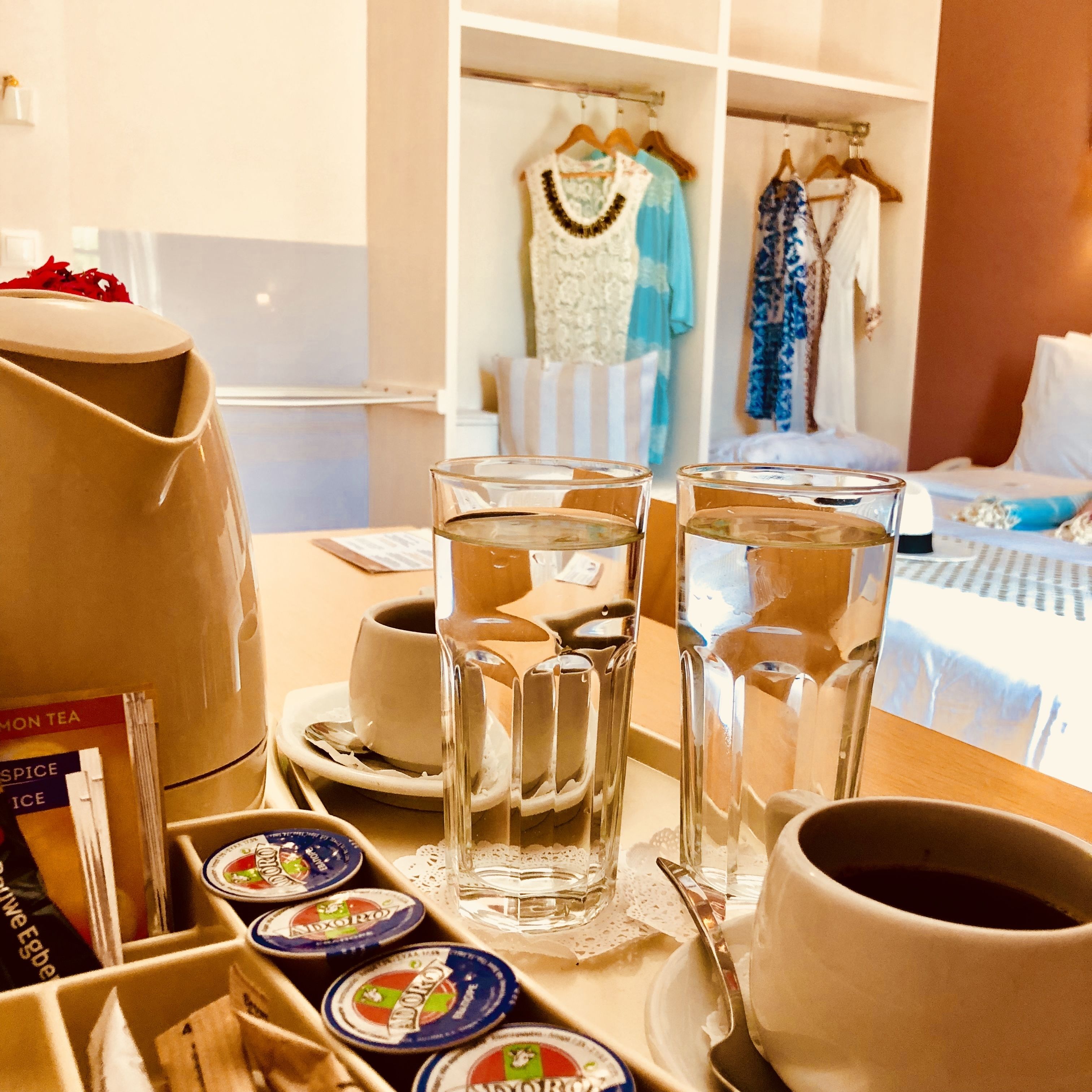 A hotel room with a dining table set with tea and coffee cups, a tray with condiments, and a wardrobe with hanging clothes.