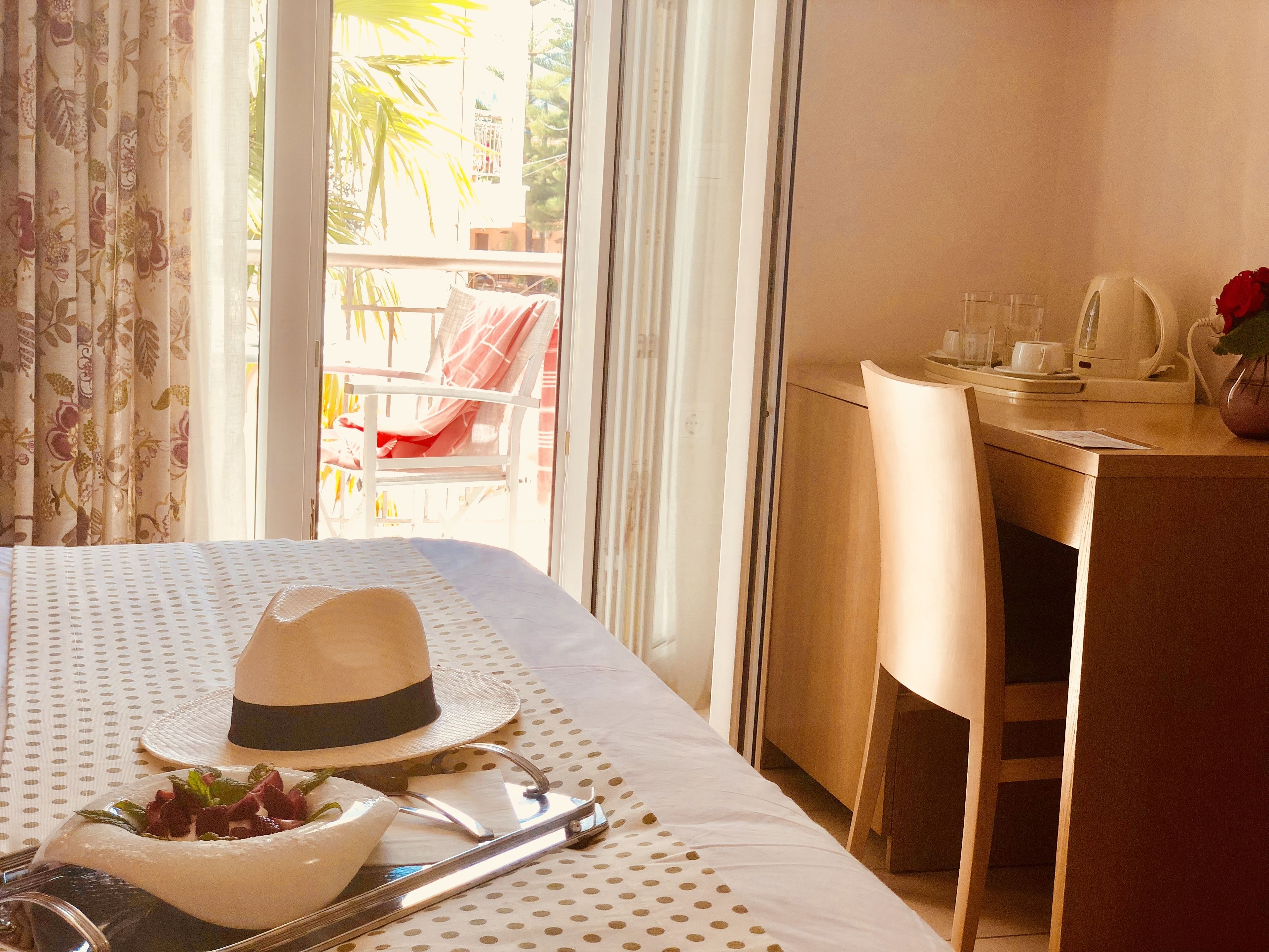 A hotel room with a bed, a dining table with a hat and a bowl of fruit, a desk with a telephone and a vase of flowers, and a balcony with a chair and a table.
