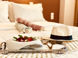 Una cama bien hecha con ropa blanca, un sombrero blanco y una bandeja con un cuenco de fruta y utensilios.