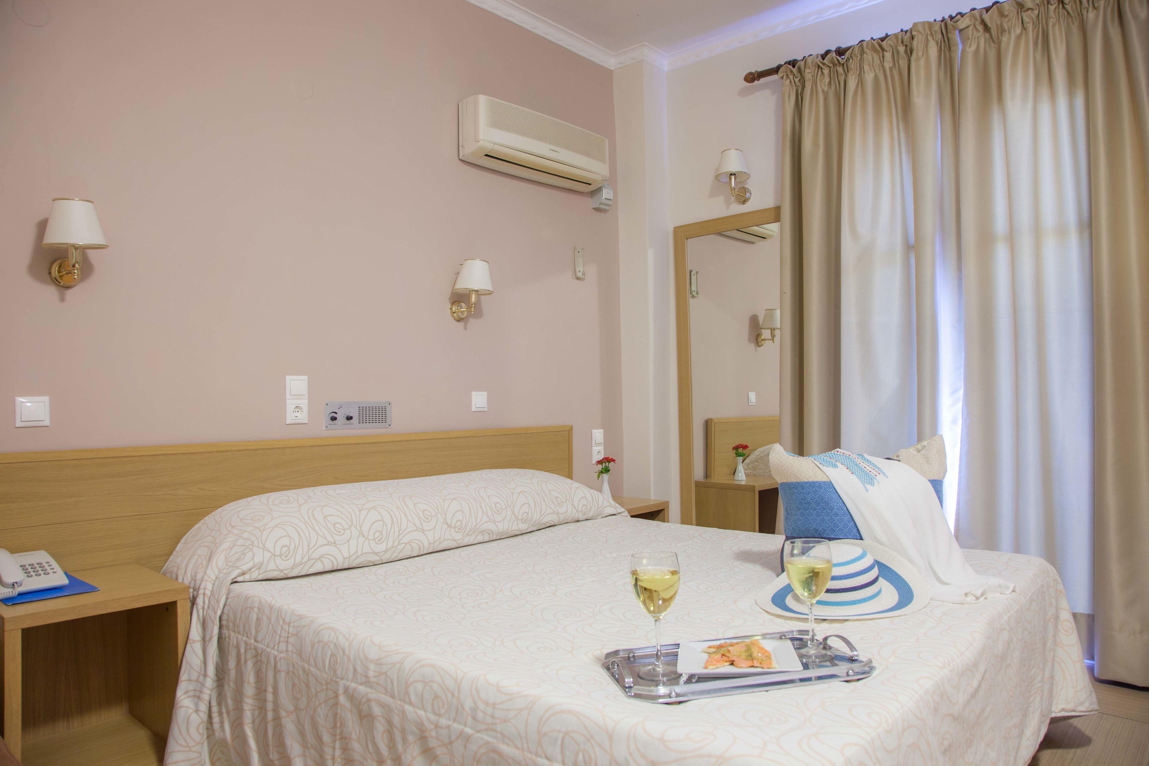 A hotel room with a bed, bedside tables, an air conditioner, a mirror, and a window with curtains.