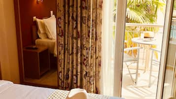 Ein Hotelzimmer mit einem Bett, einem Esstisch mit einem Hut, einem Stuhl, einem kleinen Tisch mit einer Lampe und Blick auf einen Balkon mit Pflanzen.