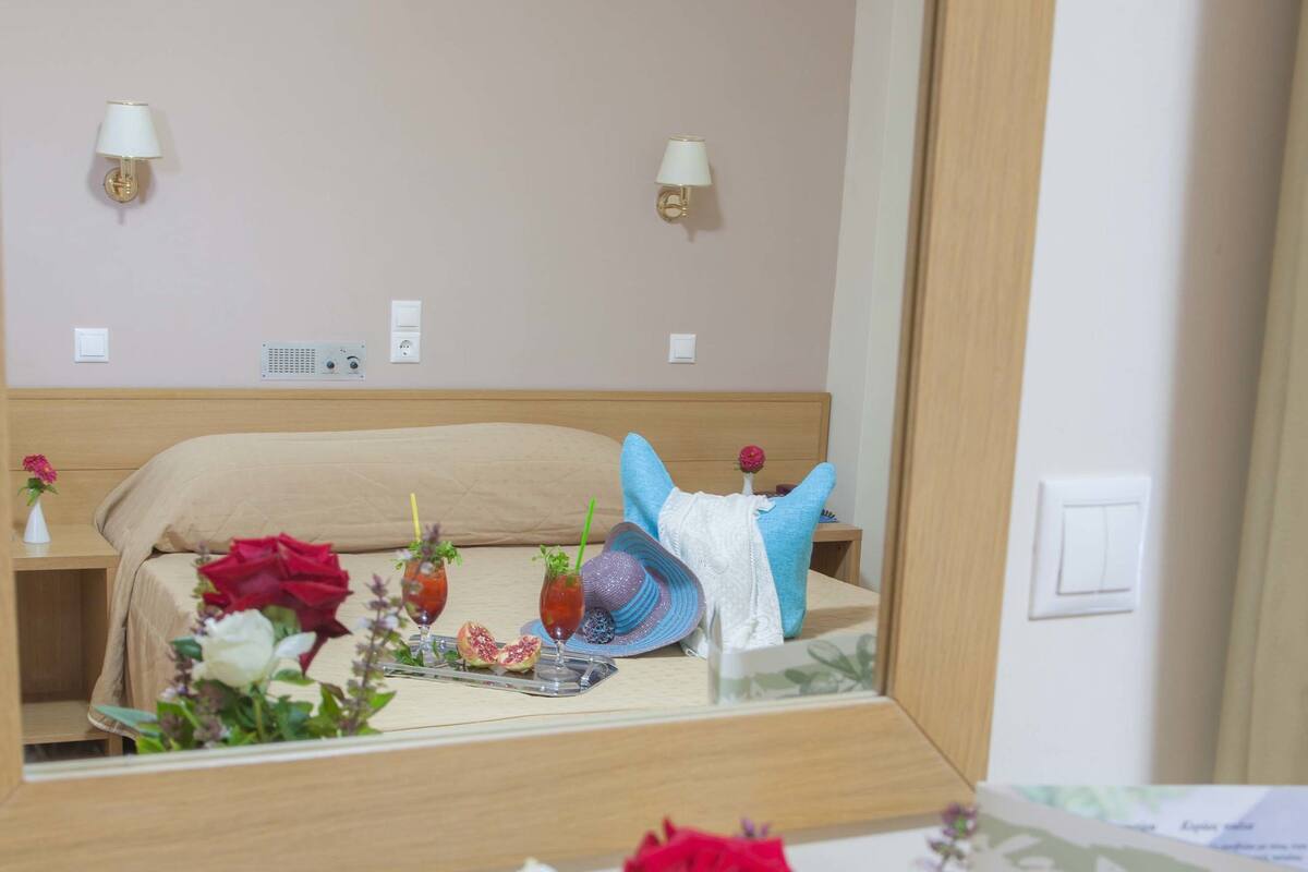 A hotel room with a bed, a bedside table with a vase of flowers, a mirror reflecting the room, and a card on the table.